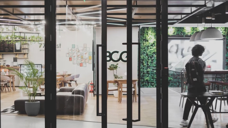 A Person with A Backpack Walking Into a Modern Office Space with Glass Doors and Indoor Plants