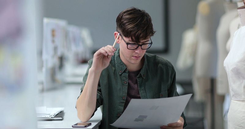 Young man in glasses reviewing a paper thoughtfully, considering how to tailor a letter of continued interest