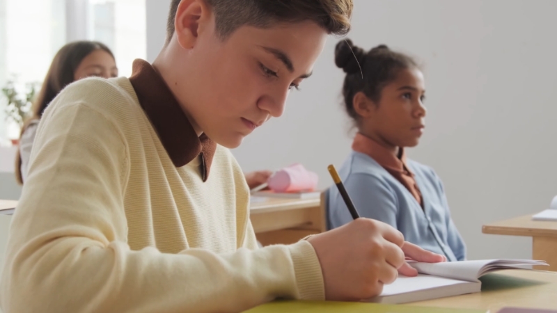 A young boy writes in a notebook while sitting in a classroom, with a female student seated behind him