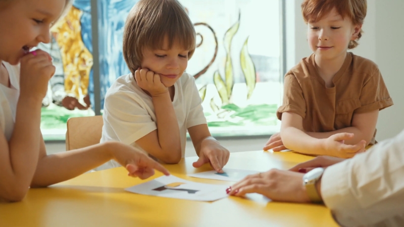 Three children sit at a table, interacting with a teacher as they point at pictures during a learning activity