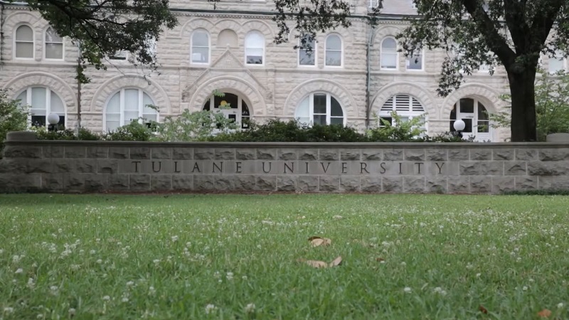 Stone wall engraved with Tulane University in front of historic campus building surrounded by grass and trees