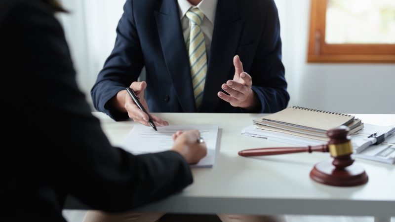 Two people in a law office reviewing legal documents across a desk with a gavel, notepad, and files visible