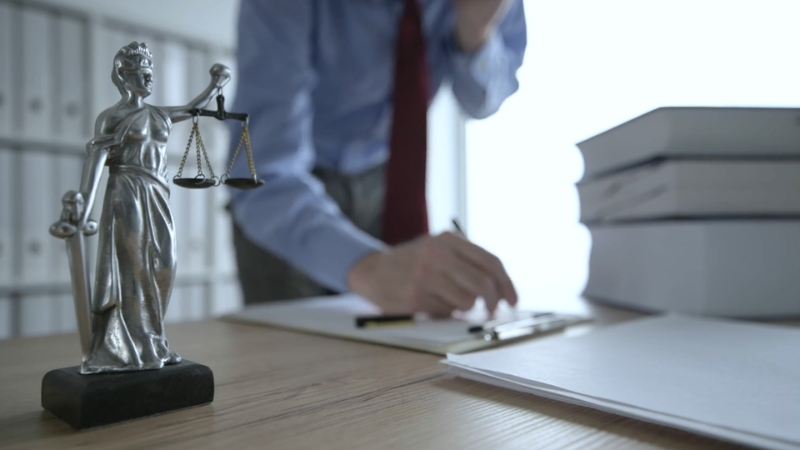 A lawyer stands at a desk writing on a clipboard beside law books and a Lady Justice statue inside a law office