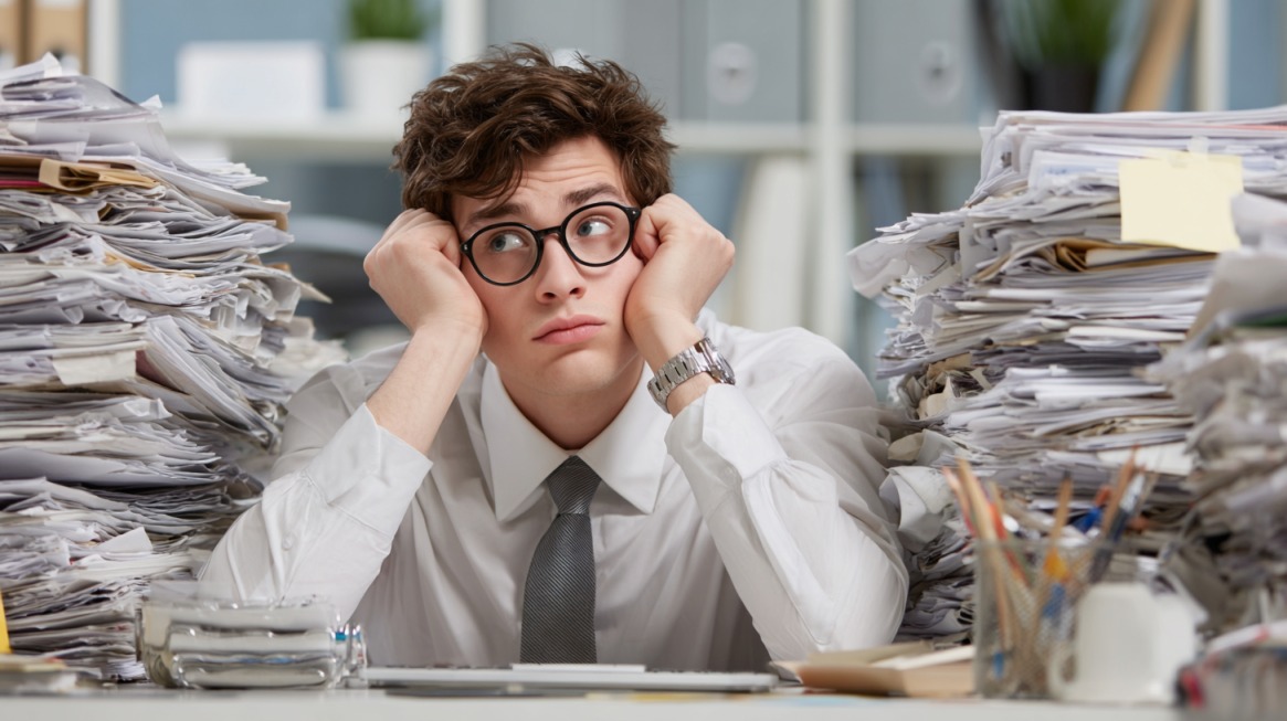 Young man in office attire looking stressed between tall stacks of paperwork, symbolizing workplace overwhelm