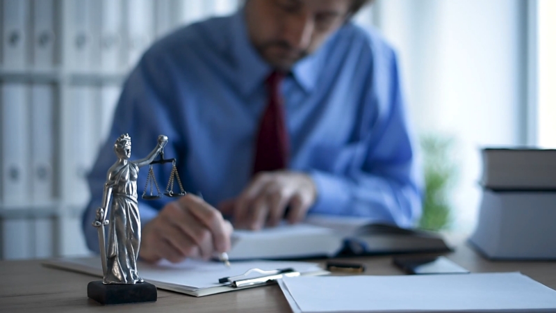 A lawyer writes at his desk beside a statue of Lady Justice and a stack of law books inside a law office