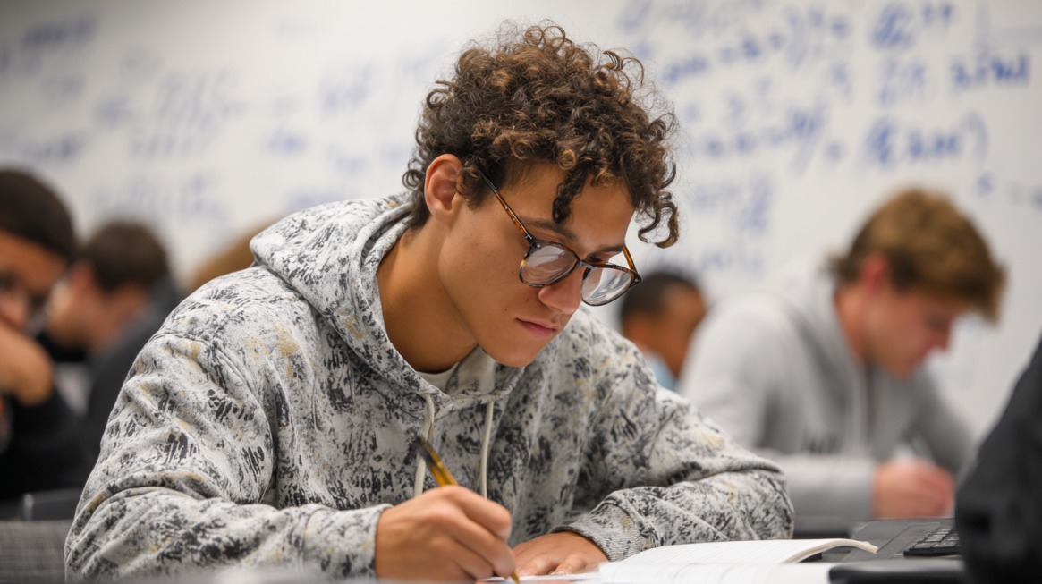 Student concentrating on algebra problems in class, writing in a notebook with equations on the whiteboard behind