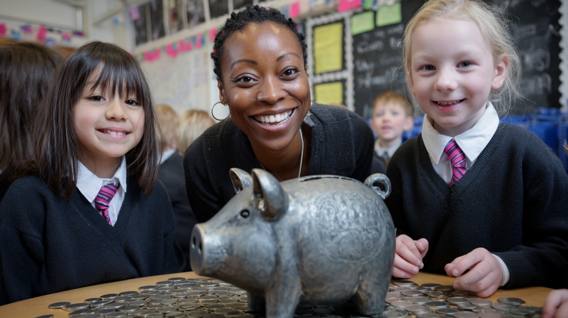 Two smiling students and a teacher gathered around a piggy bank surrounded by coins in a classroom