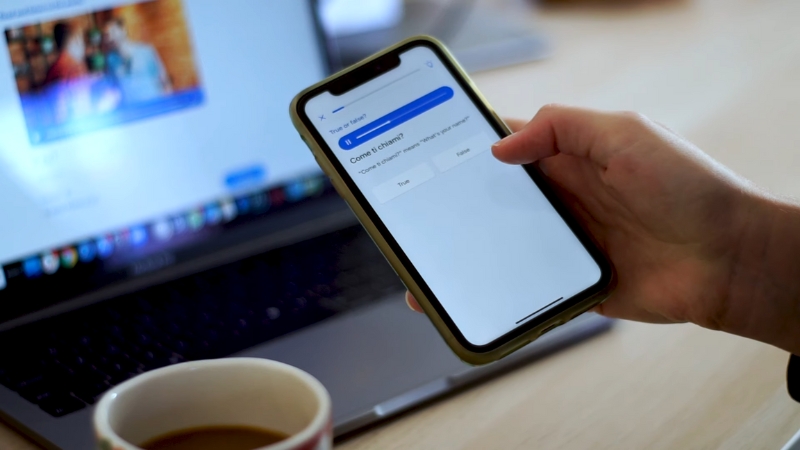 A person uses the Busuu app on a smartphone to study Italian with a laptop and coffee nearby