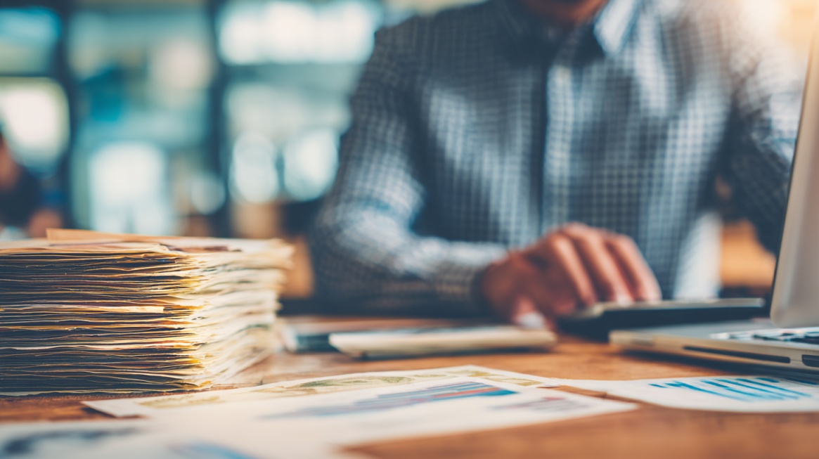 Person working on a laptop with a large stack of documents on a desk